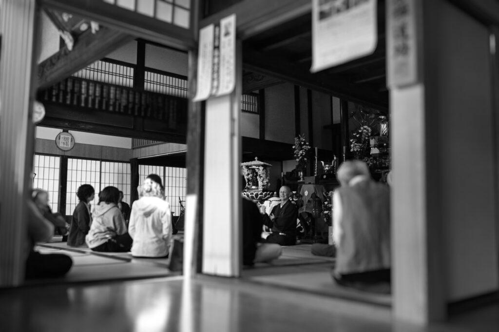 慶學寺に集まった多くの様々な年齢の方々へやさしく身振り手振りで坐禅の説明を解説するGarvey住職のようすのモノクローム画像